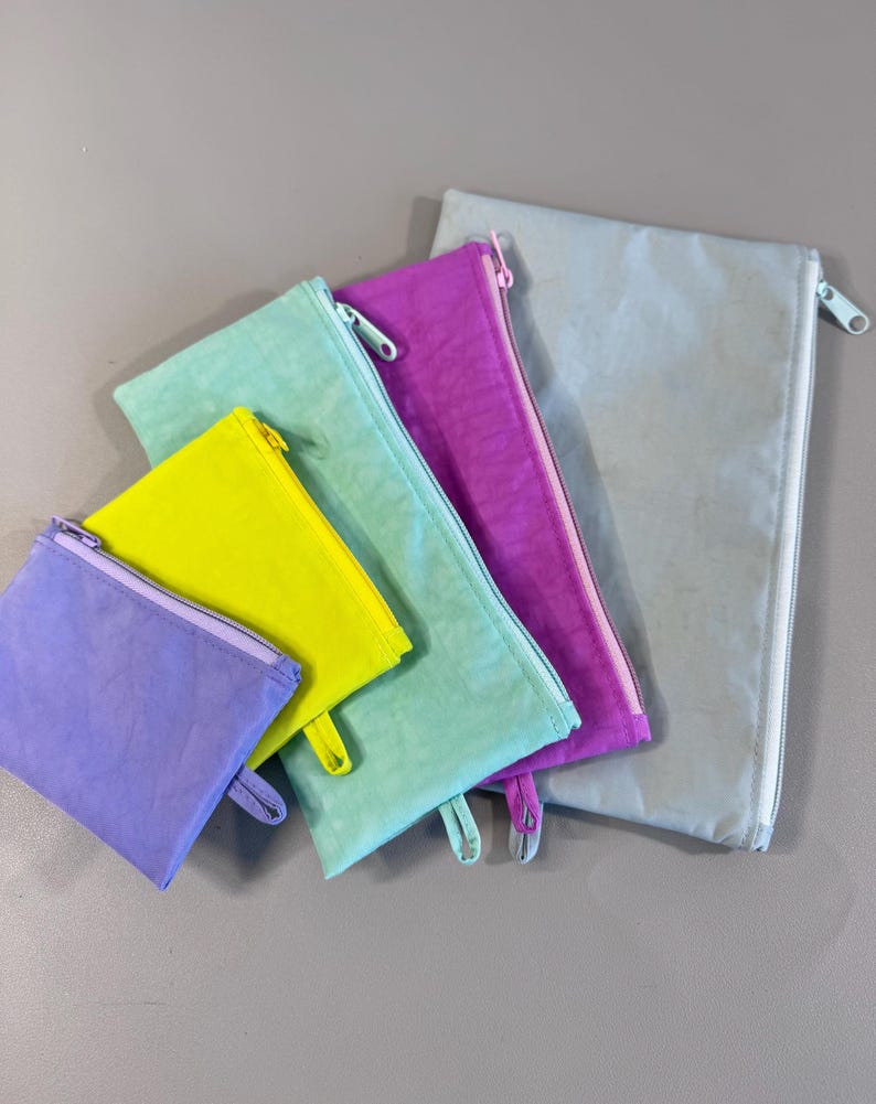 May include: A collection of colorful zippered pouches in various sizes. The pouches are in shades of purple, yellow, teal, and gray. Each pouch has a matching zipper and pull tab.