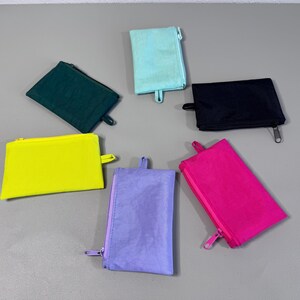 May include: A collection of six small, rectangular zippered pouches in various colors: teal, black, yellow, green, purple, and hot pink. Each pouch has a matching zipper and a small loop on one side.