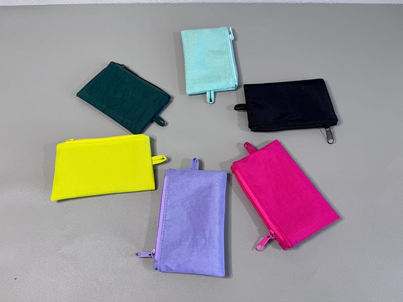 May include: A collection of six small, rectangular zippered pouches in various colors: teal, black, yellow, green, purple, and hot pink. Each pouch has a matching zipper and a small loop on one side.