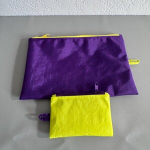 May include: Two zippered pouches in contrasting colors. A large, rectangular purple pouch with a yellow zipper and trim sits above a smaller, square yellow pouch with a purple zipper. Both have a loop on the side.