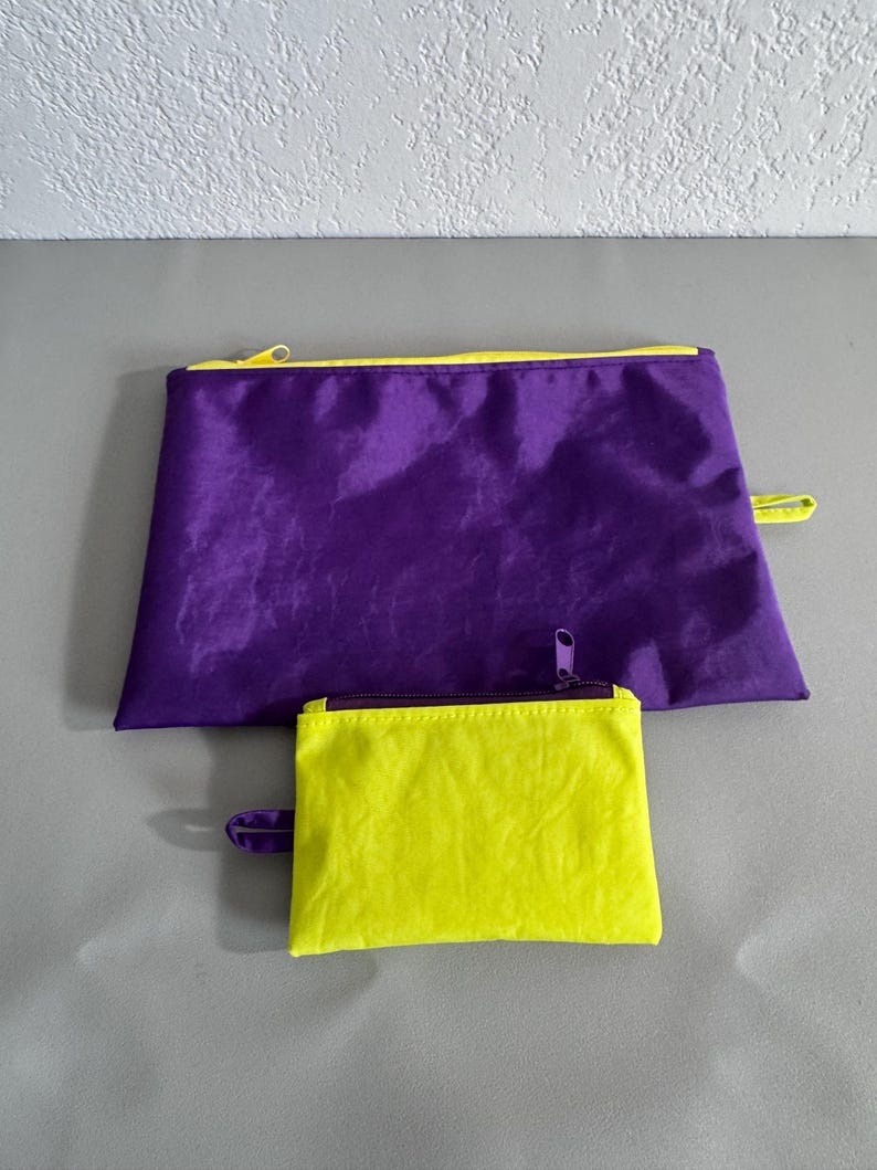 May include: Two zippered pouches in contrasting colors. A large, rectangular purple pouch with a yellow zipper and trim sits above a smaller, square yellow pouch with a purple zipper. Both have a loop on the side.