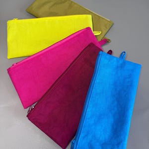 May include: A collection of colorful zippered pouches in shades of yellow, pink, blue, burgundy, and olive green. Each pouch has a matching zipper and a small loop on one end. The pouches are made of a crinkled fabric.