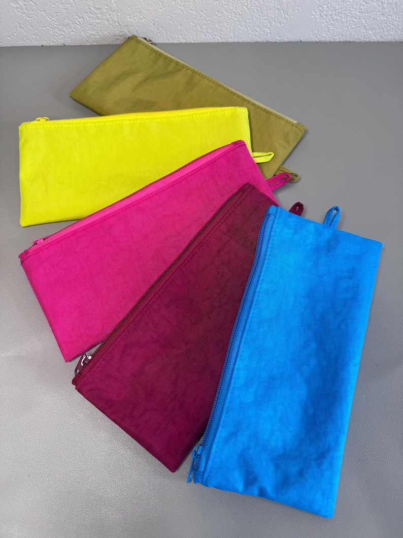 May include: A collection of colorful zippered pouches in shades of yellow, pink, blue, burgundy, and olive green. Each pouch has a matching zipper and a small loop on one end. The pouches are made of a crinkled fabric.