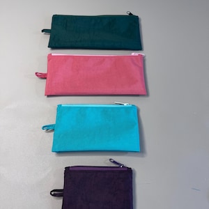 May include: Four rectangular zippered pouches in teal, pink, teal, and purple. Each pouch has a matching zipper pull and a contrasting zipper. The pouches are arranged vertically against a neutral background.