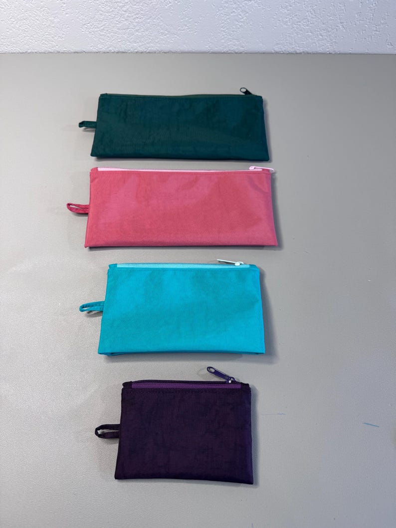 May include: Four rectangular zippered pouches in teal, pink, teal, and purple. Each pouch has a matching zipper pull and a contrasting zipper. The pouches are arranged vertically against a neutral background.