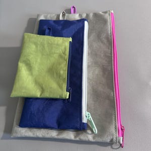May include: A set of three fabric pouches in varying sizes and colors. The largest pouch is a light tan with a pink zipper. A medium-sized pouch is blue with a white zipper. The smallest pouch is lime green.