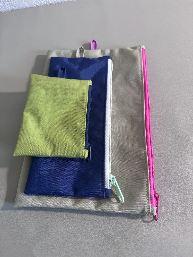 May include: A set of three fabric pouches in varying sizes and colors. The largest pouch is a light tan with a pink zipper. A medium-sized pouch is blue with a white zipper. The smallest pouch is lime green.