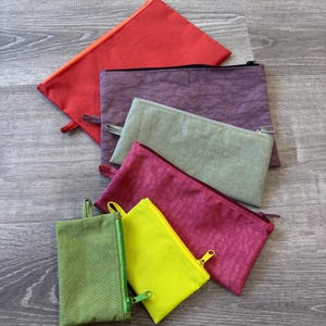 May include: A collection of colorful zippered pouches in various sizes. The pouches are in shades of red, purple, green, pink, and yellow. Each pouch has a matching zipper and a small fabric loop. The pouches are arranged on a wooden surface.