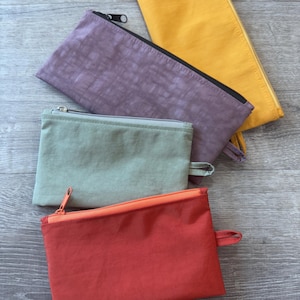 May include: Four rectangular zippered pouches in shades of red, green, purple, and yellow. Each pouch has a matching zipper and a small fabric loop on one end. The pouches are arranged on a gray wooden surface.