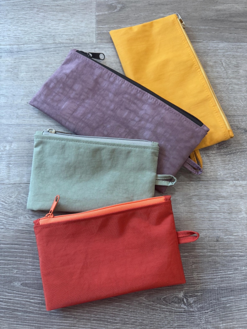May include: Four rectangular zippered pouches in shades of red, green, purple, and yellow. Each pouch has a matching zipper and a small fabric loop on one end. The pouches are arranged on a gray wooden surface.