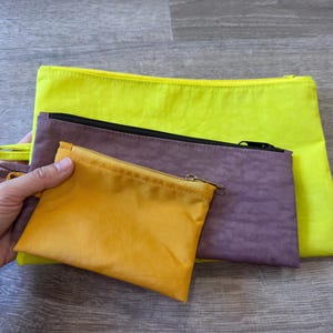 May include: A collection of colorful zippered pouches in various sizes. The pouches are in shades of yellow, purple, and gold. Each pouch has a zipper closure. The smallest pouch is held in a hand.