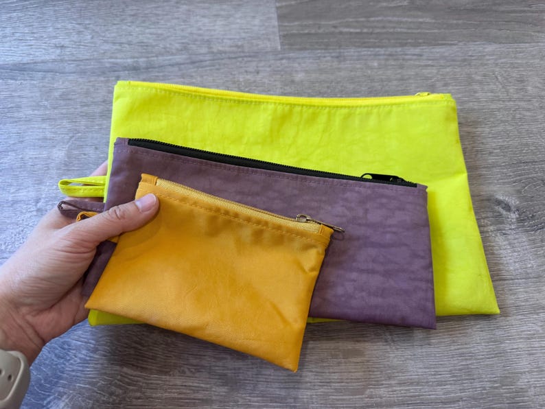 May include: A collection of colorful zippered pouches in various sizes. The pouches are in shades of yellow, purple, and gold. Each pouch has a zipper closure. The smallest pouch is held in a hand.
