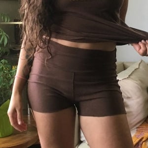May include: A woman wearing a brown short-sleeved shirt with gold wings and a heart design and the text "Sweet Skin is Near". She is also wearing brown shorts.