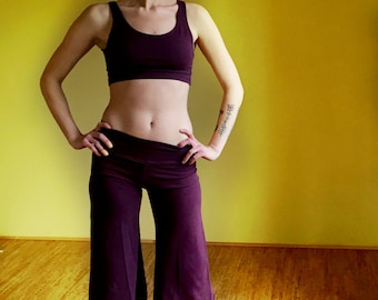 May include: A woman wearing a purple sports bra and matching wide-legged pants. She is standing in front of a yellow wall and looking to the right.