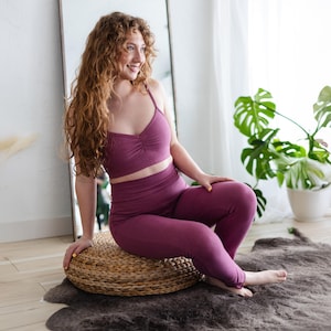 Peut inclure: Ensemble de vêtements de sport deux pièces de couleur mauve foncé. L'ensemble comprend un crop top à fines bretelles et un legging taille haute. Le modèle est assis sur un pouf rond en osier. Un grand miroir et une plante en pot sont en arrière-plan.