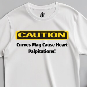 Peut inclure: T-shirt blanc avec le texte "CAUTION Curves May Cause Heart Palpitations!" en noir et jaune. Le t-shirt a un col rond et des manches courtes. Une paire de chaussures beiges est placée à droite du t-shirt.