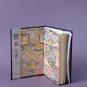 May include: An open black journal with a colorful patterned cover. The pages are blank and the journal is open to the middle.