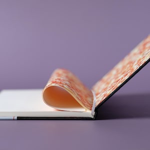 May include: A black and orange patterned notebook with a blank page open. The notebook is lying on a purple surface.