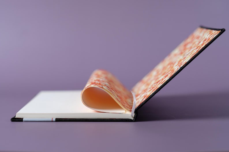 May include: A black and orange patterned notebook with a blank page open. The notebook is lying on a purple surface.