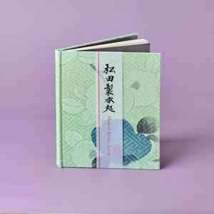 Könnte beinhalten: Ein kleines, offenes Buch mit hellgrünem Einband mit floralem Muster und japanischem Text. Ein weißes Band mit japanischem Text und den Worten "Made in Kyoto, Japan" befindet sich auf der Vorderseite. Das Buch liegt auf einer violetten Oberfläche.