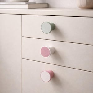 Op de afbeelding: Close-up van een crèmekleurige kast met drie lades, elk met een ronde knop. De knoppen wisselen af in kleur: groen, roze en roze, met een wit midden. De kast heeft een neutraal kleurenschema.