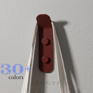 Pode incluir: Um gancho de parede vermelho escuro com topo arredondado e dois pinos circulares, segurando uma alça de tecido branco. O texto 30+ colors é exibido em azul no lado esquerdo e a palavra SAMOYA é visível na alça de tecido branco.