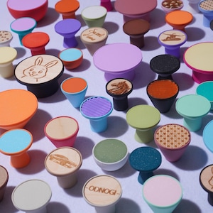 May include: A collection of colorful, decorative bottle stoppers in various shapes and sizes. Some stoppers feature engraved designs, including a rabbit, a horse, and a car. The stoppers are in shades of orange, blue, green, purple, and pink.