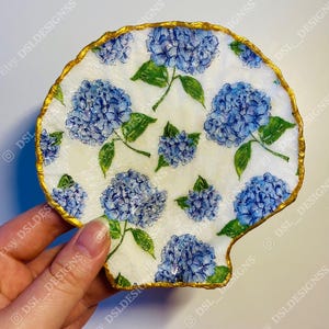 May include: A white ceramic dish shaped like a seashell with a gold rim. The dish is decorated with a blue and green floral pattern of hydrangeas.