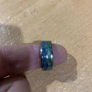 May include: A silver ring with a blue, green, and white inlay. The ring is on a finger.