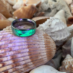 May include: A dark grey metal ring with a green and black resin inlay. The ring is sitting on a pile of seashells.