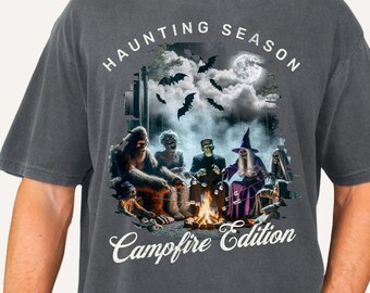 Puede incluir: Camiseta gris oscuro con un diseño temático de Halloween. El diseño presenta una escena de fogata con varios monstruos y el texto "HAUNTING SEASON Campfire Edition." La camiseta está disponible en cuatro colores.