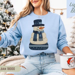 May include: Light blue sweatshirt featuring a snowman design. The snowman has a black top hat, a carrot nose, and a blue scarf. The sweatshirt has the text "Light Blue" in the upper right corner and "GILDAN 18000" at the bottom.