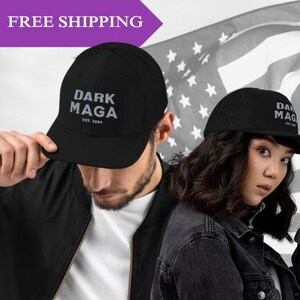 May include: Two people wearing black baseball caps with the text "DARK MAGA EST. 2024" in white. One cap is a standard baseball cap, and the other is a mesh trucker cap.