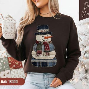 May include: A dark chocolate-coloured sweatshirt with a patchwork snowman design. The snowman has a black hat, an orange carrot nose, and a red and blue scarf. The sweatshirt has the text "GILDAN 18000" at the bottom.