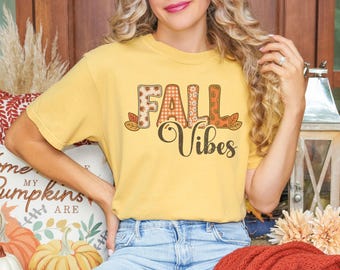 May include: A yellow t-shirt with the words "FALL Vibes" in a fall-themed design. The letters are decorated with plaid, floral, and leaf patterns. The shirt is a solid yellow color.