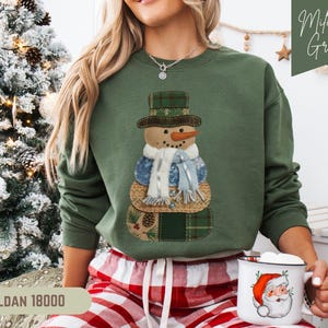 Peut inclure: Sweat-shirt vert militaire avec un motif de bonhomme de neige portant un chapeau et une écharpe à carreaux. Le bonhomme de neige a un nez en carotte et des yeux en bouton. Le sweat-shirt est un Gildan 18000.