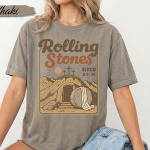 May include: Khaki-coloured t-shirt with a graphic featuring the text "Rolling Stones since A.D. 33" in brown and orange. The graphic includes a depiction of a tomb with a stone and crosses. The shirt is a Comfort Colors brand.