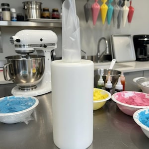 May include: A white cylindrical pastry bag holder with a clear plastic pastry bag inserted. The image shows a kitchen setting with a stand mixer, bowls of colorful frosting, and hanging pastry bags.