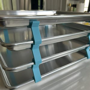 May include: A stack of four stainless steel baking sheets held together by light blue supports. The trays are rectangular with rounded edges and a shiny finish. The supports are a contrasting color, creating a functional and visually appealing kitchen storage solution.