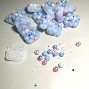 May include: A collection of small, light blue resin cubes filled with white, blue, and pink beads. The cubes are scattered on a white surface, along with loose beads in various colors.