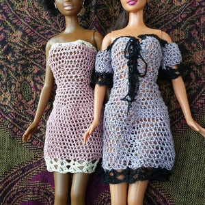 Può includere: Due bambole Barbie che indossano abiti all'uncinetto. La bambola a sinistra indossa un abito rosa con bordi bianchi. La bambola a destra indossa un abito lavanda con bordi neri.