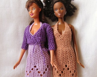 May include: Two dolls wearing crocheted dresses. The doll on the left is wearing a purple dress with a matching cardigan. The doll on the right is wearing a brown dress.