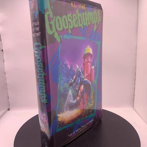 以下が含まれることがあります： R.L. Stineの「Goosebumps: Attack of the Mutant」のヴィンテージVHSテープ。カバーには、赤い塔を背景に、マントを着た不気味な姿が描かれています。タイトルは緑色で、紫とティールのボーダーがあります。