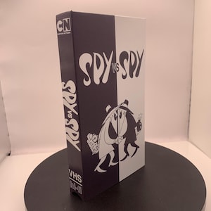 May include: A black and white VHS tape box featuring the cartoon characters Spy vs. Spy. The box has the Cartoon Network logo and the words "SPY vs SPY" in a stylized font. The box is on a black circular stand.