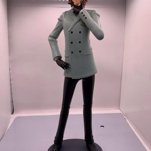 May include: A figurine of a person in a light green double-breasted coat, black pants, and black shoes. The figure is standing on a gray circular base. The figure is wearing black gloves and has brown hair.