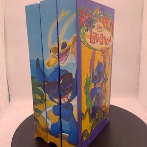 Puede incluir: Tres cajas de DVD de Lilo & Stitch. Los sets de cajas presentan a Stitch en varias poses, con el título "Lilo & Stitch" y "Vol 3" visibles. Las cajas tienen una combinación de colores azul y amarillo, con imágenes de Stitch y otros personajes.