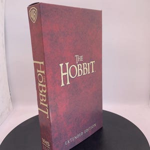 Peut inclure: Une cassette VHS de "The Hobbit: Extended Edition" dans un étui bordeaux. Le titre est en lettres dorées, avec le logo Warner Bros. en haut à gauche. La cassette VHS est debout sur une surface noire.