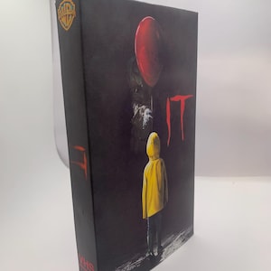 Può includere: Una custodia per videocassetta VHS nera del film IT, che mostra un bambino con un impermeabile giallo davanti a una figura scura che tiene un palloncino rosso. Il titolo IT è stampato in rosso sul fronte e sul lato, con il logo Warner Bros.