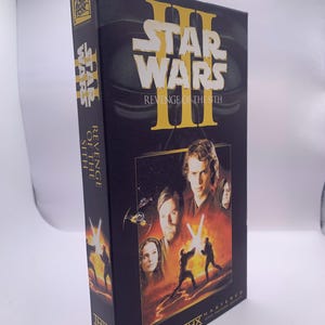 Op de afbeelding: Een zwarte VHS-cassettedoos voor Star Wars Episode III Revenge of the Sith. De hoes bevat gele tekst, een portret van Anakin Skywalker, Obi-Wan Kenobi en Padmé Amidala, en een scène van twee figuren die duelleren met lichtzwaarden. THX-logo inbegrepen.