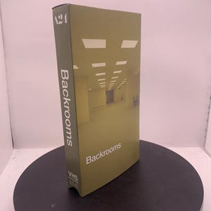 Peut inclure: Un boîtier de cassette VHS rectangulaire avec un fond jaune moutarde présentant une image d'un long couloir vide avec des plafonniers fluorescents. Le texte A24, Backrooms et VHS est imprimé en blanc sur le devant et le côté de l'emballage.